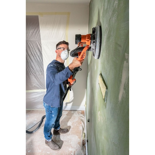 Persoon schuurt een muur met een BLACK+DECKER schuurmachine. Draagt masker en bril.