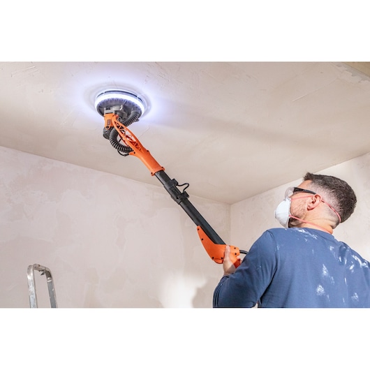 Persoon schuurt plafond met BLACK+DECKER wandenschuurmachine met LED-licht, draagt beschermende kleding.