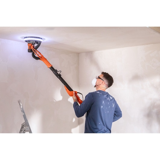 Persoon schuurt plafond met BLACK+DECKER BEDW700A8S gipsmuurschuurmachine. Draagt masker en bril.
