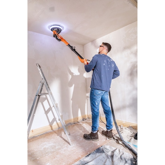 Persoon schuurt plafond met BLACK+DECKER gipsplaten schuurmachine met LED, ladder en afdekzeil erbij.