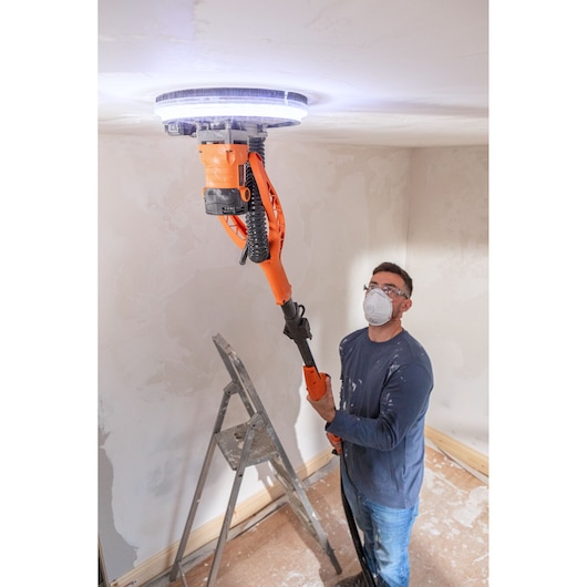 Persoon schuurt plafond met BLACK+DECKER gipsplaat schuurmachine met LED-licht in een bouwruimte.