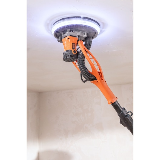 BLACKDECKER plafondschuurmachine met LED-licht schuurt een witte plafond. Oranje handvat en slang zichtbaar.