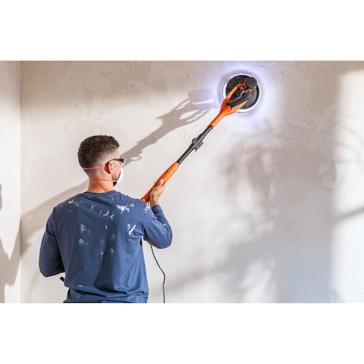 Persoon schuurt muur met een oranje BLACK+DECKER schuurmachine. Draagt masker en veiligheidsbril.