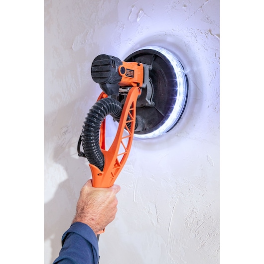 Hand gebruikt een BLACK+DECKER muurschuurmachine met LED-licht op een witte structuurmuur.