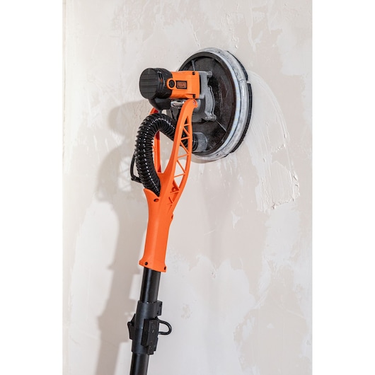 Een BLACK+DECKER gipsmuurschuurmachine met oranje handvat schuurt een muur. Rond schuurhoofd zichtbaar.