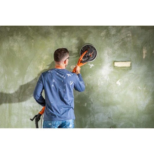 Persoon schuurt groene muur met BLACK+DECKER wandenschuurmachine tijdens renovatie, tool met beide handen vast.