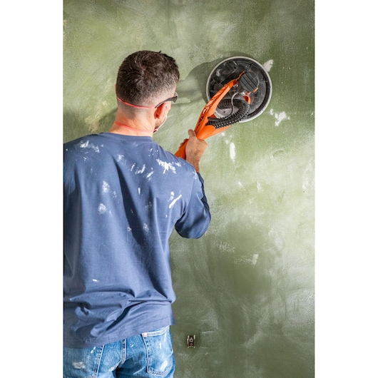 Persoon schuurt een muur met een BLACK+DECKER schuurmachine, draagt veiligheidsbril en werkkleding.