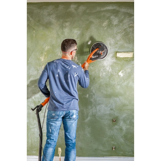 Persoon schuurt een muur met een oranje BLACK+DECKER schuurmachine, draagt een bril en casual kleding.