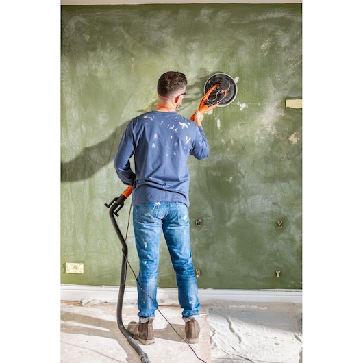 Persoon schuurt een groene muur met een BLACKDECKER wandenschuurmachine, gekleed in jeans en blauw shirt.