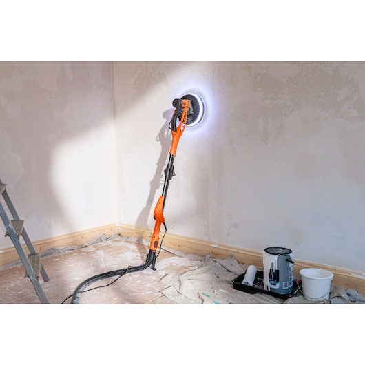 BLACK+DECKER muurschuurmachine met LED-licht schuurt muur, verfbenodigdheden en doek op de vloer.