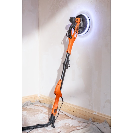 Oranje BLACK+DECKER gipsmuurschuurmachine met LED-licht schuurt een muur in een hoek.