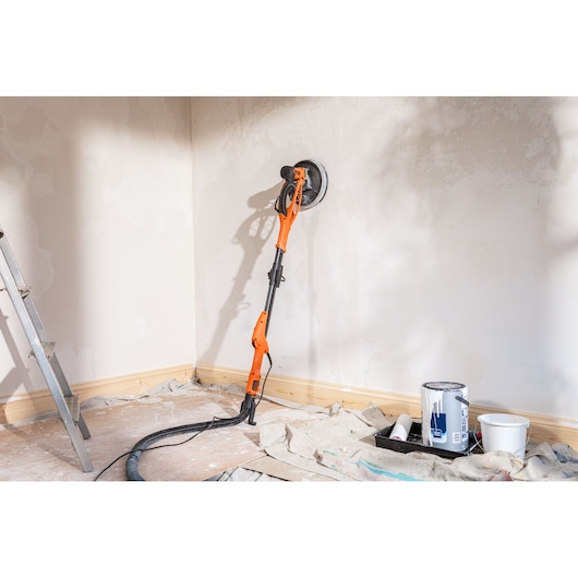 BLACK+DECKER wandenschuurmachine op een gepleisterde muur. Ladder, verf en gereedschap op de vloer.