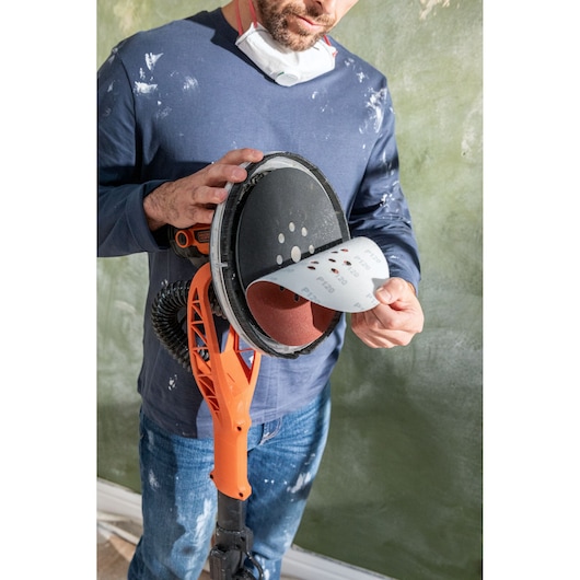Persoon vervangt schuurpapier op een oranje BLACK+DECKER wandenschuurmachine. Mondmasker en werkkleding zichtbaar.