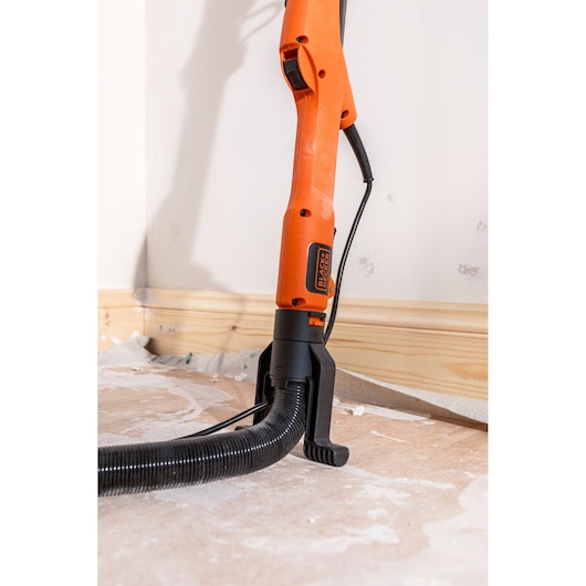 BLACK+DECKER oranje wandschuurmachine met slang op een vloer bij een muur, gebruikt voor renovatie.