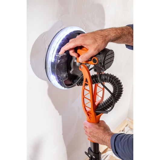 Een persoon gebruikt een BLACK+DECKER wandenschuurmachine met LED-licht op pleisterwerk.