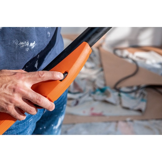 Persoon drukt op de aan-knop van een oranje BLACK+DECKER gereedschap, rommelige werkplek op de achtergrond.