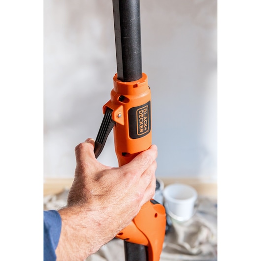 Hand verstelt oranje BLACK+DECKER stokgereedschap in kamer met schilderbenodigdheden op de achtergrond.