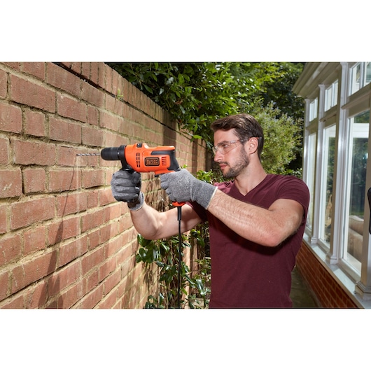 BLACK+DECKER BEH200 perceuse filaire utilisée à l’extérieur sur un mur en briques, utilisateur avec des gants.