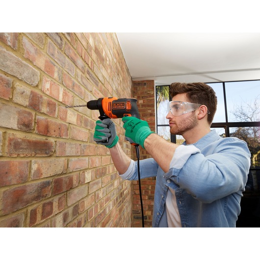 Persoon gebruikt BLACK+DECKER BEH710 snoerboormachine op bakstenen muur binnen, met handschoenen.