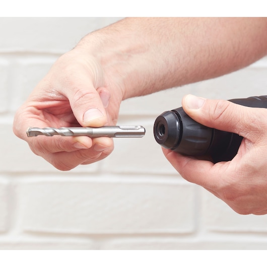 Photo de mains tenant une perceuse BLACKDECKER BEHS01K et un foret métallique devant un mur en briques blanches.