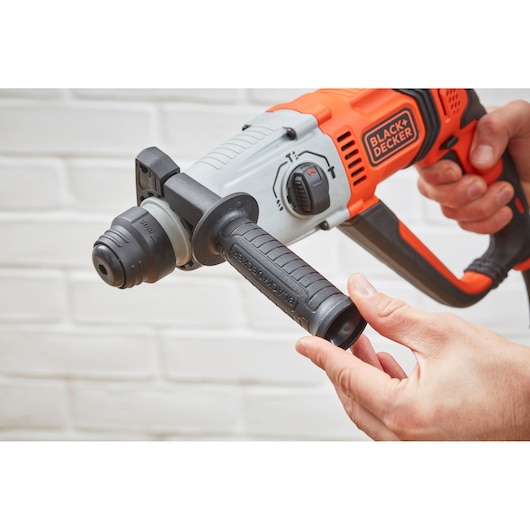 Photo de la perceuse rotative BLACK+DECKER BEHS02K avec poignée orange et noire, tenue en main devant un mur de briques blanches.