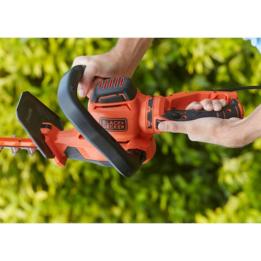 BLACK+DECKER BEHTS551 heggenschaar vastgehouden met twee handen, oranje en zwart, tuin op de achtergrond.