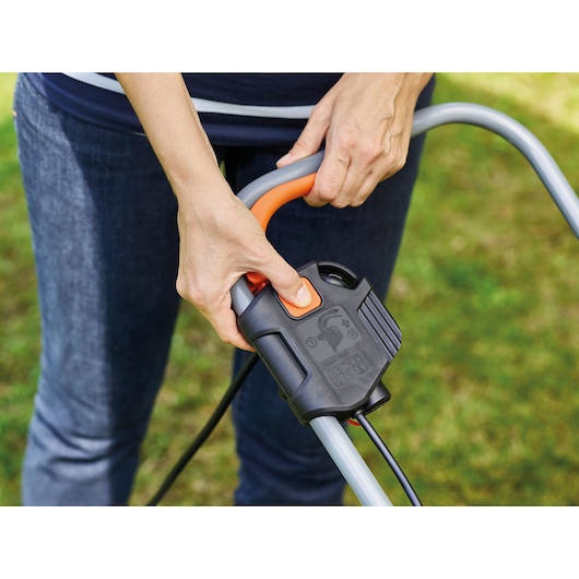 Photo en gros plan d’une personne appuyant sur le bouton de démarrage de la poignée de la tondeuse BLACK+DECKER BEMW351GL2 en extérieur.