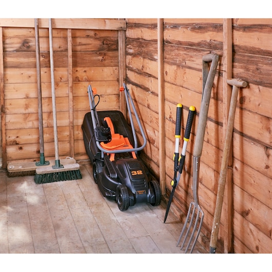 Tondeuse BLACKDECKER BEMW351 avec poignée orange rangée dans un abri en bois, entourée d’outils de jardin.