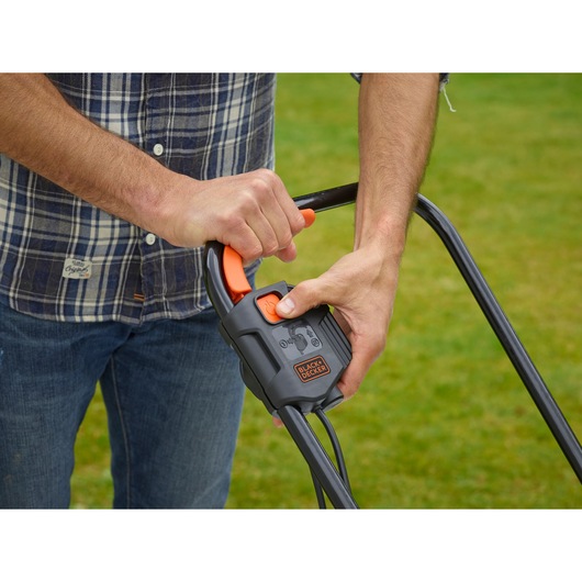 Gros plan sur des mains utilisant la poignée de tondeuse BLACK+DECKER BEMW451 avec levier de commande sur gazon.