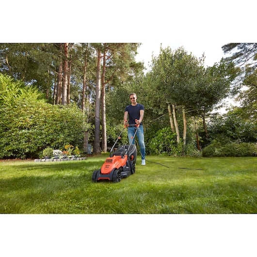 Photo de la tondeuse électrique orange BLACK+DECKER BEMW461BH en utilisation sur une pelouse verte entourée d’arbres et de plantes.