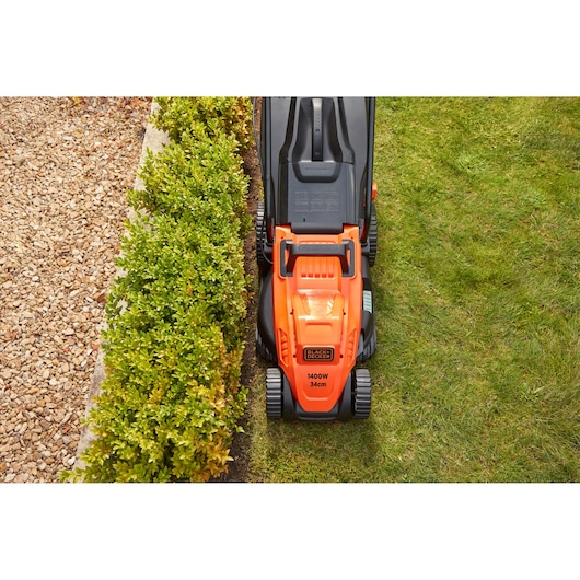 Tondeuse BLACK+DECKER BEMW461BH orange et noire tondant l’herbe, vue du dessus, près d’une haie et d’un chemin de gravier.