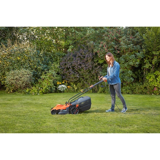 Persoon gebruikt de BLACKDECKER BEMW461ES elektrische grasmaaier in oranje en zwart op het gras in de tuin.