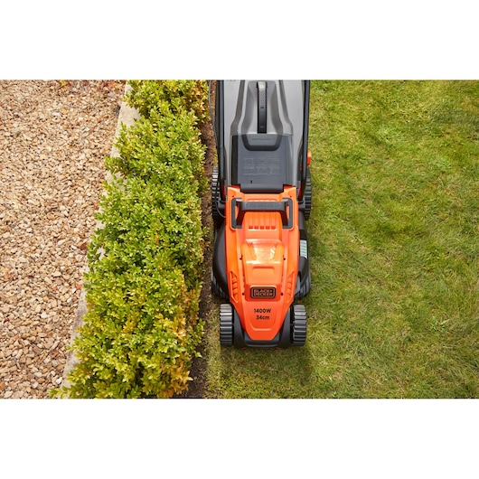BLACKDECKER BEMW461ES oranje grasmaaier bovenaanzicht op gras naast haag en grindpad.