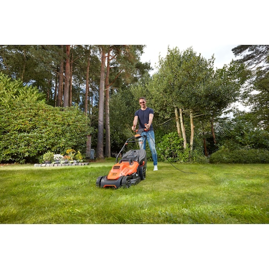 Foto van BLACKDECKER BEMW471ES grasmaaier in gebruik op gras in een tuin met hoge bomen op de achtergrond.