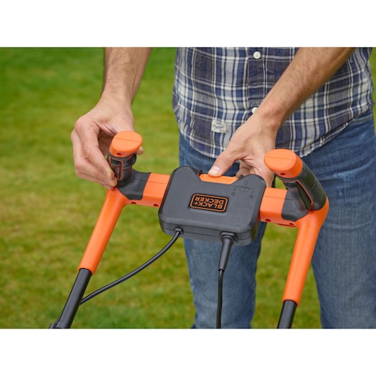 Photo en gros plan de la poignée de tondeuse BLACK+DECKER BEMW481BH avec commandes orange et noires en extérieur.