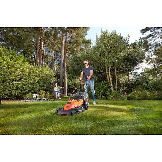BLACKDECKER BEMW481ES grasmaaier in gebruik op gras in een tuin met bomen en een kind op de achtergrond.