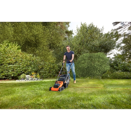 Photo de la tondeuse électrique BLACK+DECKER BEMWP400-QS utilisée sur une pelouse verte avec des plantes de jardin en arrière-plan.
