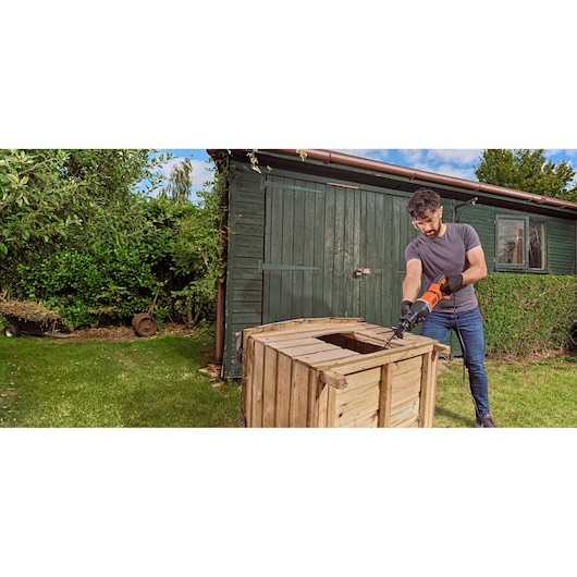 Een persoon gebruikt een BLACK+DECKER reciprozaag (model BES301) om een houten krat buiten bij een groene schuur te zagen. De persoon draagt handschoenen en werkt op het gras.