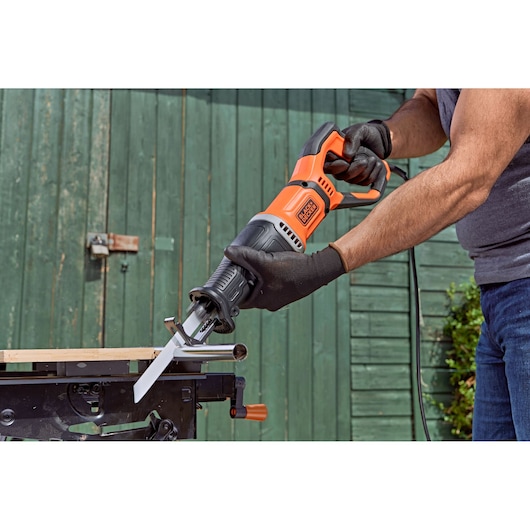 Een persoon met handschoenen gebruikt een BLACK+DECKER BES301 reciprozaag om een metalen pijp te zagen, waarbij de zaag buiten op een werkbank is geplaatst.