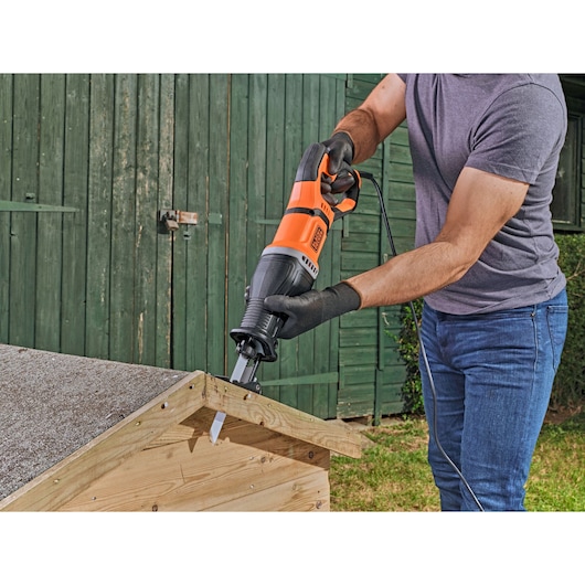 Een persoon gebruikt een BLACK+DECKER BES301 reciprozaag om een stuk hout buiten te zagen, draagt handschoenen en houdt de zaag op een houten constructie.