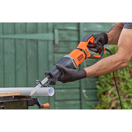 Een persoon met zwarte handschoenen gebruikt een BLACK+DECKER reciprozaag om een witte plastic pijp buiten te zagen.