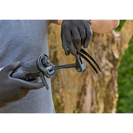 Een persoon met zwarte handschoenen houdt een BLACK+DECKER reciprozaag met een accessoire vast, buiten, met een vervaagde boomstam op de achtergrond.