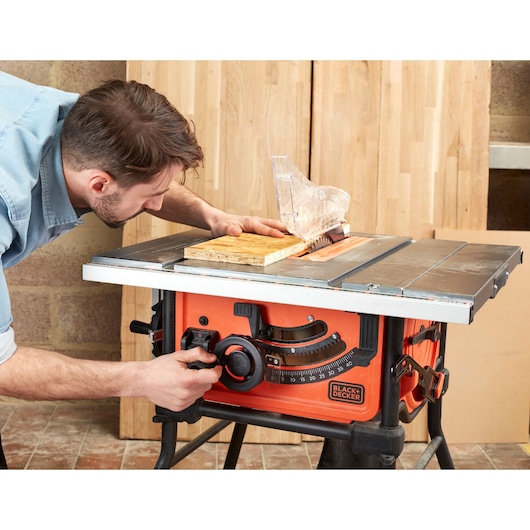 Foto van BLACK+DECKER BES720 tafelcirkelzaag in gebruik, oranje behuizing, doorzichtige zaagbladbescherming, hout wordt gezaagd.