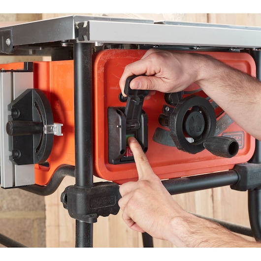Close-up van handen die de BLACK+DECKER BES720 tafelzaag aan-uitschakelaar bedienen, oranje behuizing en zwarte bediening.
