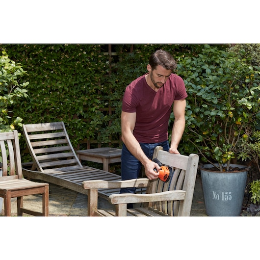 Foto van persoon die houten tuinstoel schuurt met BLACK+DECKER BEW200K detailschuurmachine in tuin.