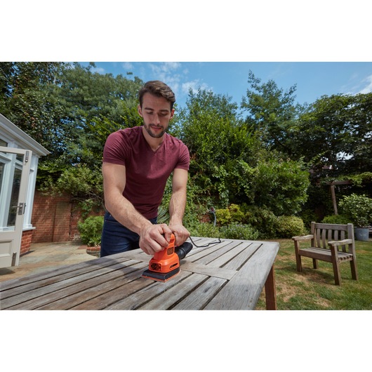 Persoon gebruikt BLACKDECKER BEW200LSA vlakschuurmachine op houten buitentafel in tuin.