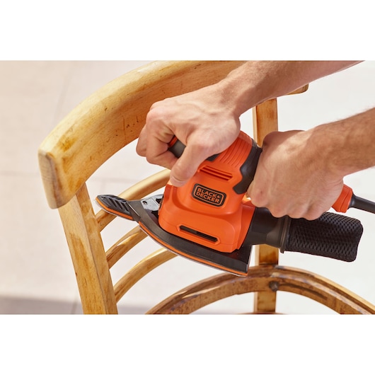 Foto van BLACK+DECKER BEW200LSA schuurmachine gebruikt op houten stoel, oranje en zwarte uitvoering.