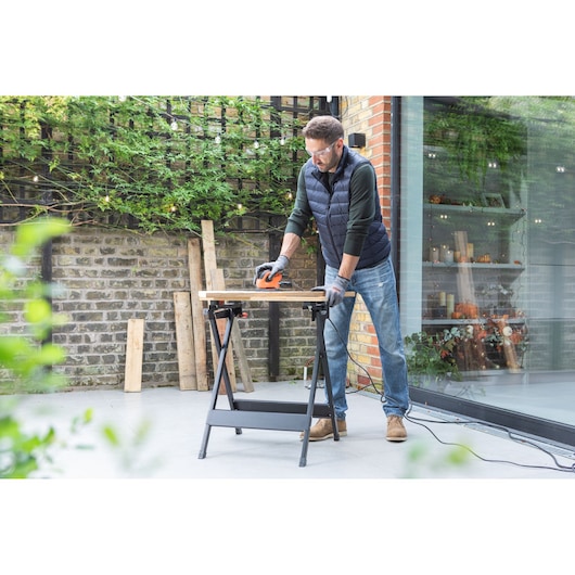 Persoon gebruikt BLACKDECKER BEW230 schuurmachine op houten plank buiten met werkbank.