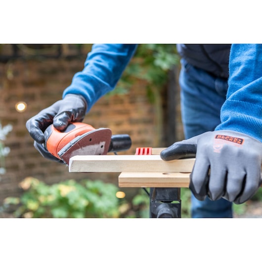 BLACKDECKER BEW230 schuurmachine gebruikt op hout, persoon met BLACKDECKER handschoenen buiten.