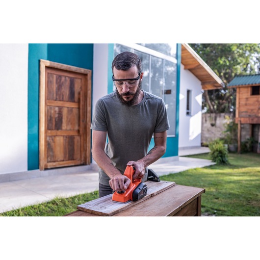 Photographie d'une personne utilisant un rabot électrique BLACK+DECKER BEW712 sur une planche en bois à l'extérieur.